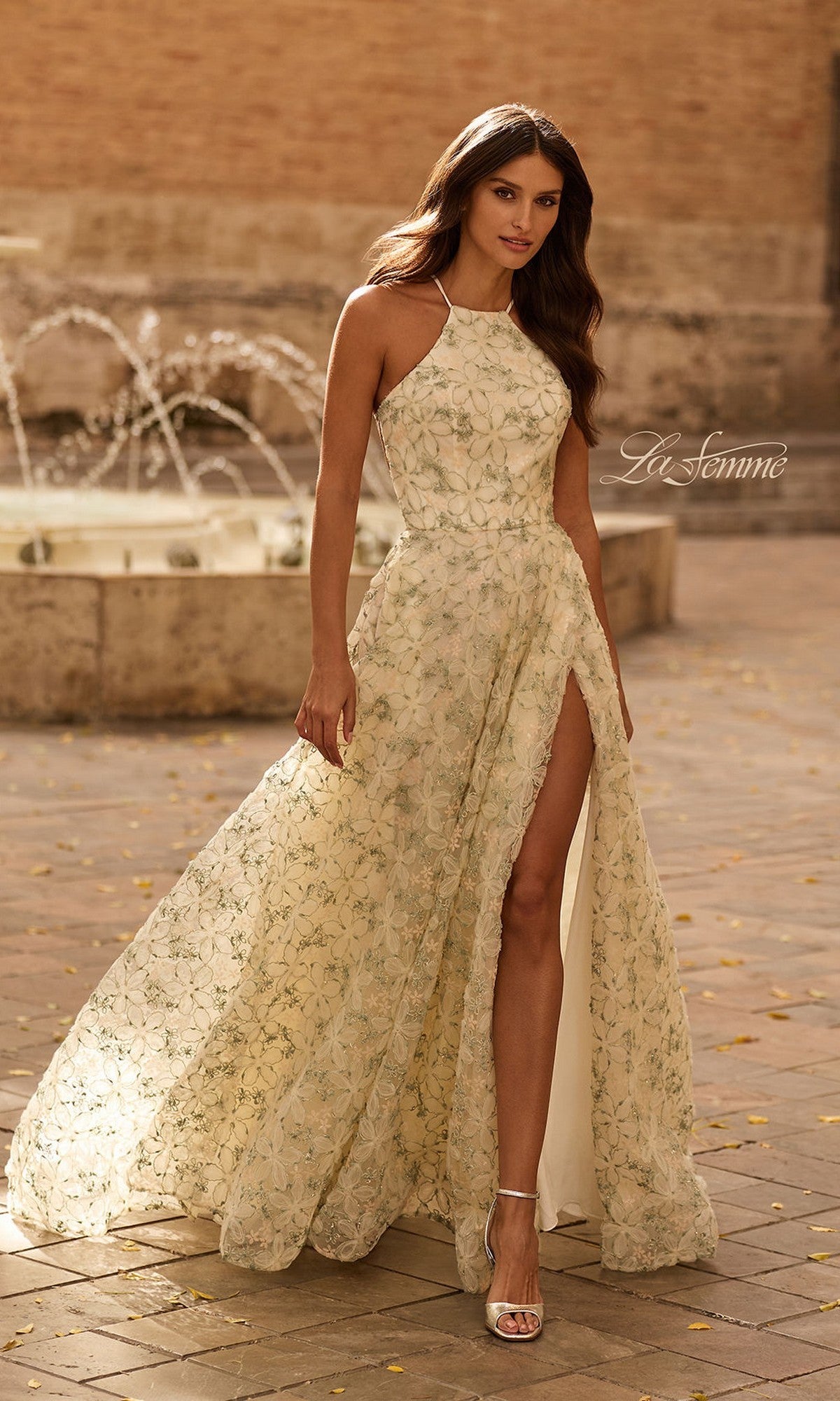 A woman stands outdoors near a stone fountain wearing the La Femme 33865 long multi-colored prom dress, featuring a sleeveless A-line design with a high leg slit and delicate floral lace embroidery, paired with silver heeled sandals.